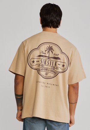 Man wearing beige t-shirt with large vintage-style "SikSilk Beach Club & Lounge" logo featuring a palm tree and text on the back.