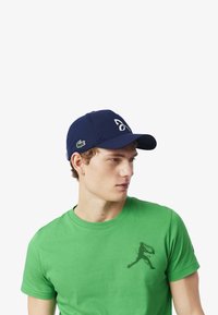 Tummansininen aluslakki, jossa on valkoinen logo ja vihreä brodeerattu alligaattori sivulla; käytetään vihreän t-paidan kanssa.