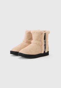 Beige suède enkelboots met een fluffy voering, ronde neus en zwarte rubberen zool. Voorzien van een zijrits met branding op een stoffen accent.