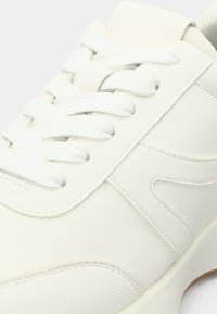 Λευκό δερμάτινο αθλητικό παπούτσι με υφασμάτινη υφή, στρογγυλή μύτη και λευκά κορδόνια. Η πλευρά διαθέτει διακριτικό ανάγλυφο σχέδιο και αντίθετη ραφή.