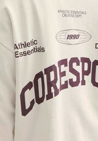 Hvid bomulds sweatshirt med "CORESPO" i store maroon bogstaver. Anden tekst inkluderer "Athletic Essentials" i sort. Glat tekstur.