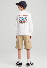 T-shirt blanc à manches longues avec un design coloré de Keith Haring au dos, associé à un short cargo beige et des baskets noires.