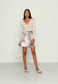 Mulher vestindo um cardigan curto bege com decote em V, saia mini de padrão animal, sandálias brancas e segurando uma pequena bolsa branca, contra um fundo simples.