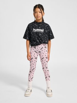 Junges Mädchen trägt ein schwarzes Hummel-Logo-T-Shirt, pinke Hummel-Logo-Leggings, weiße Socken und Turnschuhe, steht mit entspannten Armen.
