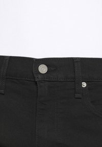 Los jeans de mezclilla negros presentan un botón de metal y detalles de remaches, con costuras visibles a lo largo de la cintura y los bolsillos laterales.