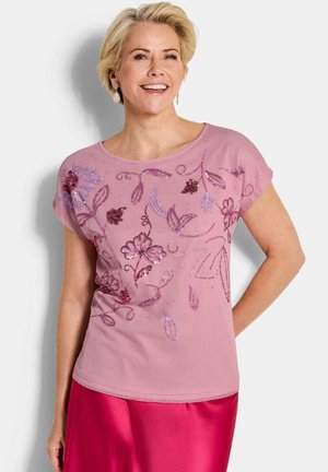 Lächelnde Frau mit kurzem blondem Haar trägt ein pinkfarbenes Oberteil mit floralen Paillettenmustern und einen knallpinken Satinstock, steht vor hellem Hintergrund.