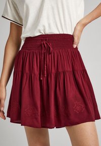 Kvinna klädd i en röd lagerkjol med smockad midja och knytning, tillsammans med en vit t-shirt med mörka kanter på ärmarna.