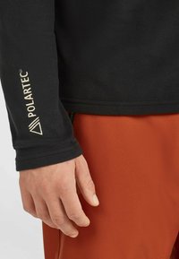 Sort fleece trøje med tommelhuller og "Polartec" logo i cremefarve, parret med orange stof nedenunder. Teksturen ser blød og varm ud.