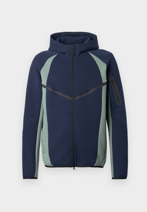 Jūras zila un gaiši zaļa ar rāvējslēdzēju aizdara jaka ar strukturētu dizainu, ar sānu kabatām, kapuci un kontrastējošiem melniem akcentiem gar šuvēm.