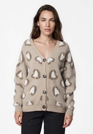 Femme aux longs cheveux bruns porte un cardigan beige avec des taches abstraites blanches et marron et de gros boutons, debout devant un fond uni.