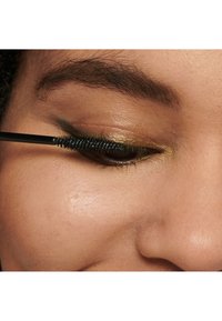 Mascara noire appliquée sur les cils à l'aide d'une brosse à picots, mettant en valeur un fard à paupières doré chaud et une texture de peau lisse.
