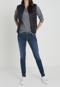 Gilet imbottito nero sopra una maglietta a maniche lunghe grigia, abbinato a jeans skinny blu e sneaker bianche. Il gilet presenta una chiusura con zip e una texture trapuntata.