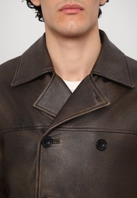 Homme portant un manteau en cuir brun foncé à double boutonnage avec de larges revers et des boutons noirs, visible de la moitié de la poitrine jusqu’aux lèvres.