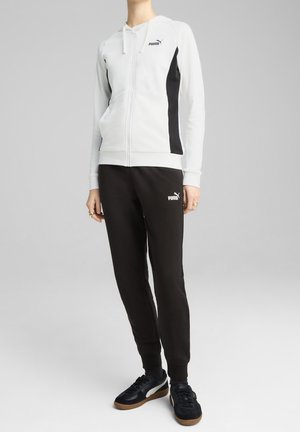 Osoba ubrana w białą bluzę z kapturem na zamek błyskawiczny z czarnymi panelami po bokach, czarne spodnie dresowe Puma i czarne trampki z gumowymi podeszwami.