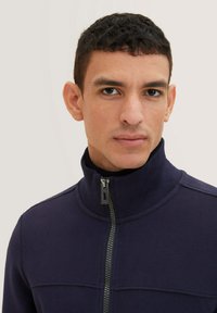 Giacca blu scuro con zip e colletto alto, caratterizzata da una zip visibile e pannelli testurizzati. Tessuto liscio con un design aderente.