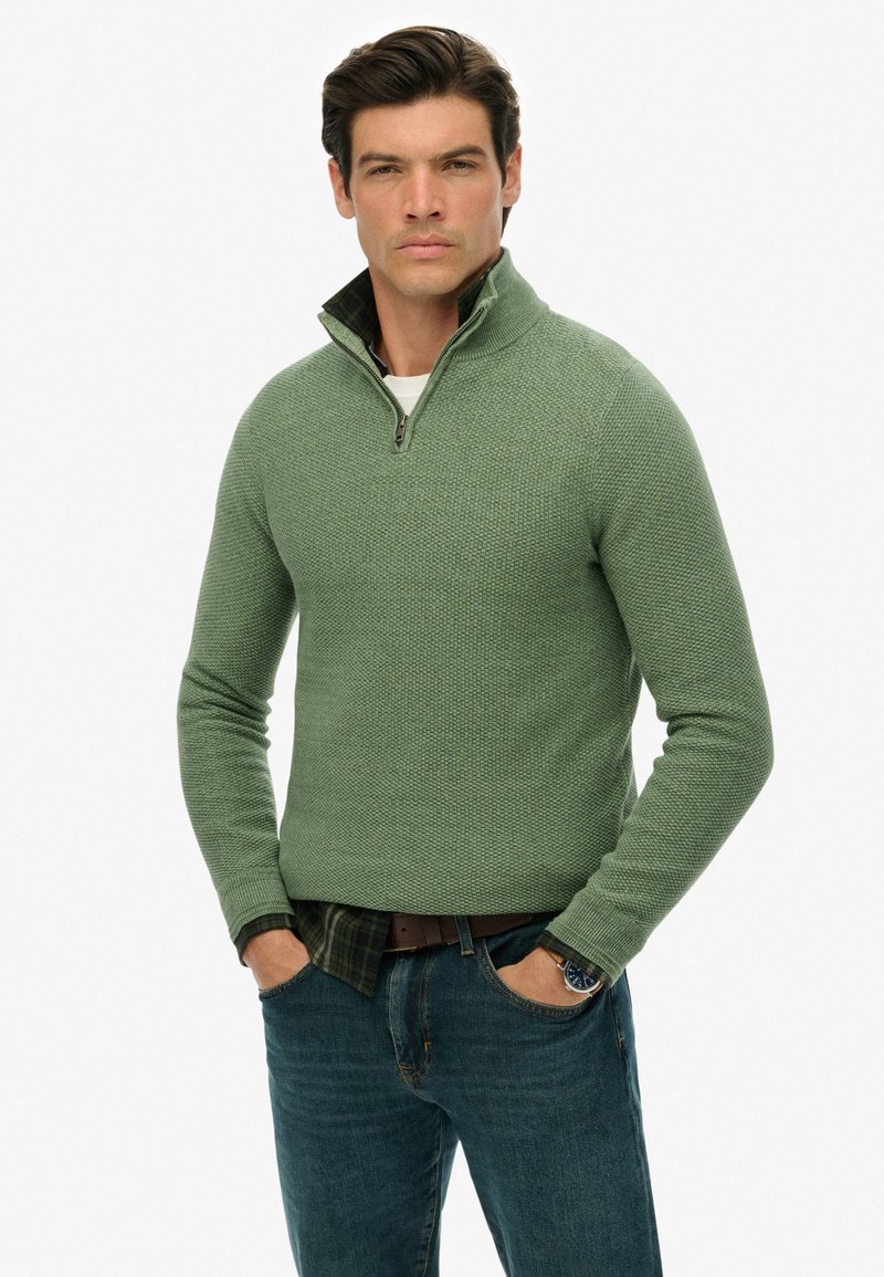 Grüner strukturierter Pullover mit einem Halbzip-Kragen, über einem dunklen karierten Hemd getragen, kombiniert mit dunklen Jeans und einer Uhr am Handgelenk.