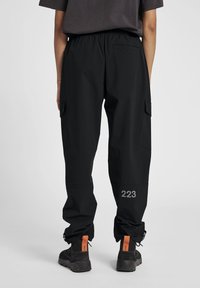 Zwarte cargobroek met een elastische tailleband, zijzakken en verstelbare boorden. Heeft een grijze "223" bedrukking nabij de zoom.