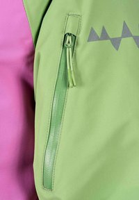 Lähikuva vihreast takin hihasta, jossa on pystysuora vetoketjullinen tasku, vihreät vetoketjun vedin sekä osa violetista vaatteesta alla.