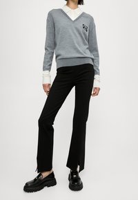 Vrouw draagt een lichtgrijze V-hals trui over een wit shirt, zwarte rechte broek met voorpanelen, en stevige zwarte loafers tegen een witte achtergrond.