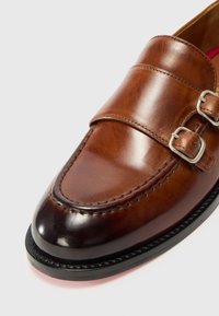 Brown leather loafer with two metal buckles, smooth texture, rounded toe, and contrasting black rubber sole.