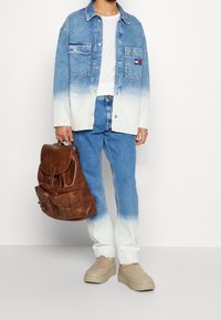 Muž oblečený v džínové bundě s modro-bílým ombré, džínách, bílém tričku a béžových platformových botách, držící hnědou koženou tašku na boku.