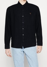 Camisa negra de manga larga con cuello tipo button-down, hecha de material texturizado. Presenta un pequeño logotipo bordado en el pecho y cierre de botones.