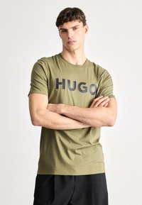 Olivovozelené bavlnené tričko s nápisom "HUGO" v tučných čiernych písmach cez prsia. Krátke rukávy a okrúhly výstrih, hladká textúra.