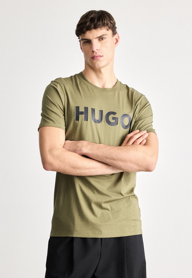Olivovozelené bavlnené tričko s nápisom "HUGO" v tučných čiernych písmach cez prsia. Krátke rukávy a okrúhly výstrih, hladká textúra.