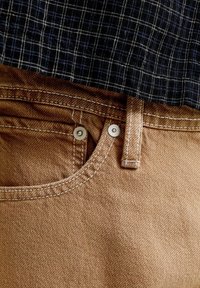 Κοντινό σε τσέπη παντελονιού από καφέ τζιν και θηλιά ζώνης με ορατό ράψιμο, σε συνδυασμό με μια μαύρη και λευκή καρό πουκάμισο που έχει μπει μέσα.