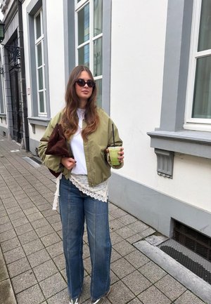 Junge Frau mit langen Haaren, die Sonnenbrille trägt, grüne Jacke, weißes Top, blaue Jeans, hält ein grünes Getränk und eine braune Tasche auf einem Bürgersteig in der Stadt.