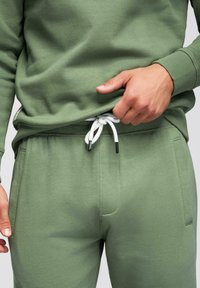 Personne portant un pantalon de survêtement vert olive et un sweat-shirt assorti, ajustant le cordon blanc de la taille du pantalon.