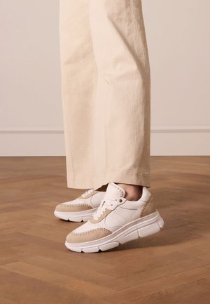 Sneakers featuring a white leather upper with beige suede accents, thick white soles, and a stitched design along the toe cap.
