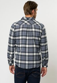 Mees nähtud tagantpoolt, seljas sinise, valge ja oranži ruudulise mustriga piklike varrukatega särk ja tumelised sinised teksad, taust on lihtne.