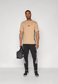 Smėlio spalvos sportiniai marškinėliai su juodais šonais ir logotipu, derinami su juodomis sportinėmis kelnėmis su baltomis vertikaliomis juostelėmis, papildyti juodais sportbačiais.