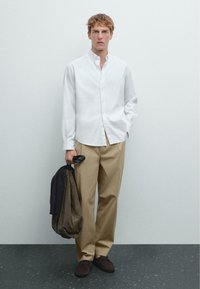 Chemise blanche à manches longues avec des boutons, pantalon beige, chaussures brunes, tenant un sac sombre, debout sur un sol sombre texturé devant un fond clair.