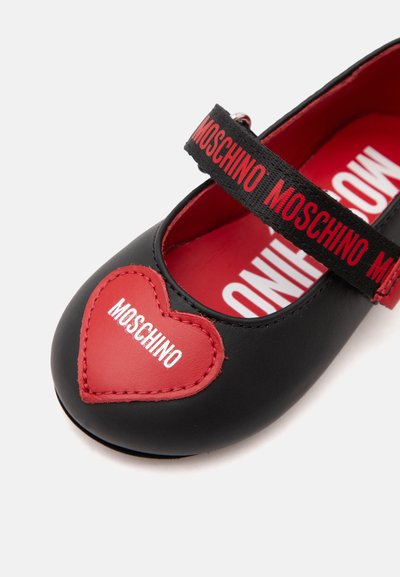 Chaussure Mary Jane noire avec un cœur rouge sur l'orteil et une bride rouge, toutes deux avec le nom de la marque Moschino en texte blanc et rouge.