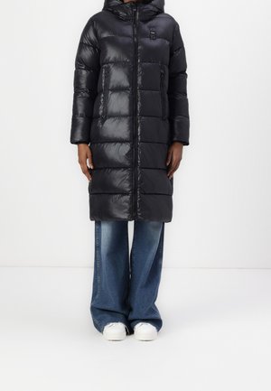 Czarna długa pikowana kurtka z kapturem, zapinana na zamek i z zasuwanymi kieszeniami po bokach, noszona na szerokich niebieskich dżinsach i białych trampkach.