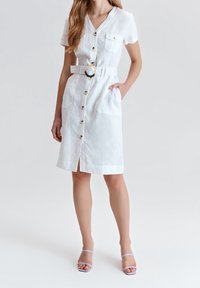 Weißes Leinenhemdkleid mit kurzen Ärmeln, Knopfleiste, zwei Taschen, Taillengürtel und einen Schlitz am Saum. Kombiniert mit blassen lila Sandalen.