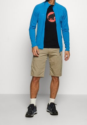 Uomo che indossa una giacca blu con cerniera sopra una maglietta nera con logo di elefante rosso, pantaloncini beige, calzini bianchi e scarpe da ginnastica nere, in piedi.