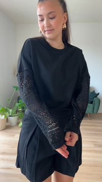 Top nero con maniche in pizzo intricate, caratterizzato da una vestibilità rilassata e polsini a coste. Abbinato a una gonna nera, ambientato in un interno luminoso e moderno.