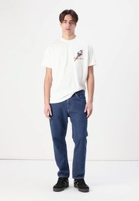 Hvid bomulds t-shirt med grafisk design på brystet, parret med blå denim jeans og sorte sneakers, stående mod en ensfarvet baggrund.