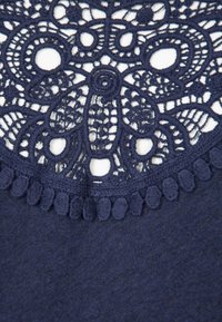 Marineblauwe stof met ingewikkelde kantdetails bij de halslijn, met cirkelvormige patronen en kleine, gewatteerde franjes.