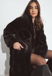 Svart fuskpälsrock med en lång, oversized design; ihop med en liten svart satinhandväska. Genomskinliga svarta strumpbyxor fulländar looken.