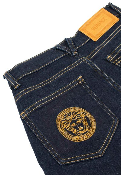 Jeans in denim scuro con ricami dorati raffiguranti un logo a testa di Medusa sulla tasca posteriore, completati da un'etichetta in pelle marrone del marchio Versace.
