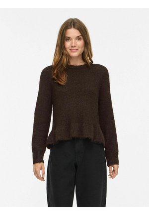 Kvinde med brunt hår iført en mørkebrun, lodden sweater med flæsekant og sorte bukser, stående foran en ensfarvet hvid baggrund.