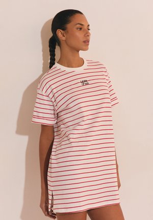 Donna con capelli intrecciati che indossa un vestito bianco a maniche corte con sottili strisce orizzontali rosse e la scritta "Sea Sun" sul petto.