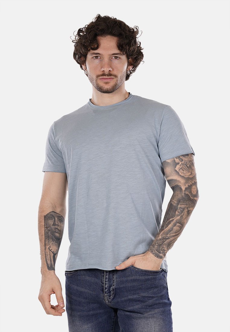 T-shirt azzurro chiaro a maniche corte con colletto rotondo, realizzato in un tessuto morbido e dal taglio rilassato. Indossato con jeans scuri; tatuaggio visibile sul braccio.