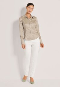 Langärmlige, hellbeige Satinbluse mit Knopfleiste und spitzem Kragen, kombiniert mit weißen, eng anliegenden Jeans und grünen Wildleder-Pumps.