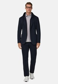 Laivastonsininen puku hupullisella takilla, kahdella etutaskulla, yhdistettynä vaaleanharmaaseen poolopaitaan ja tummiin housuihin, viimeistelty valkoisilla lenkkareilla.