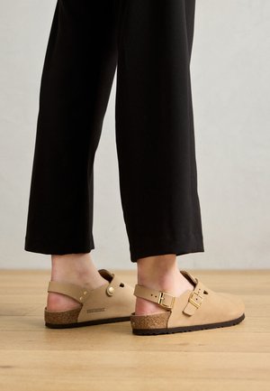 Beige Birkenstock dřeváky s korkovými stélkami, nastavitelnými přezkami a bočními výřezy, v kombinaci s černými, volně střiženými kalhotami na dřevěné podlaze.